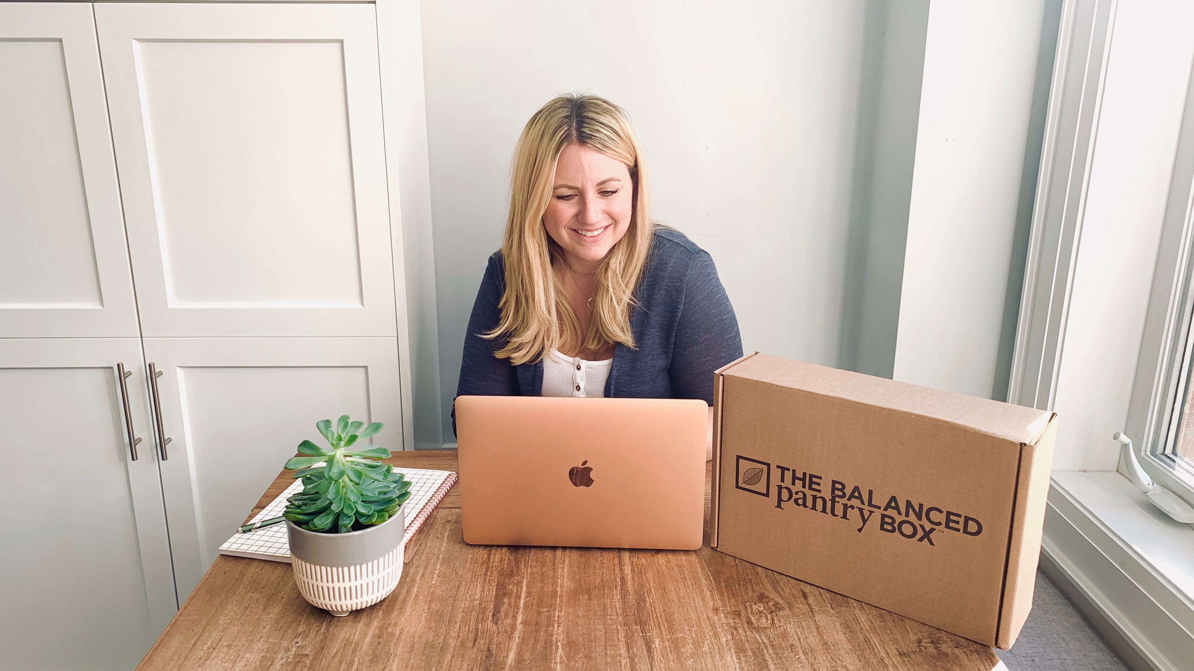 Woman using a laptop with a 'The Balanced Pantry Box' on a wooden table.
